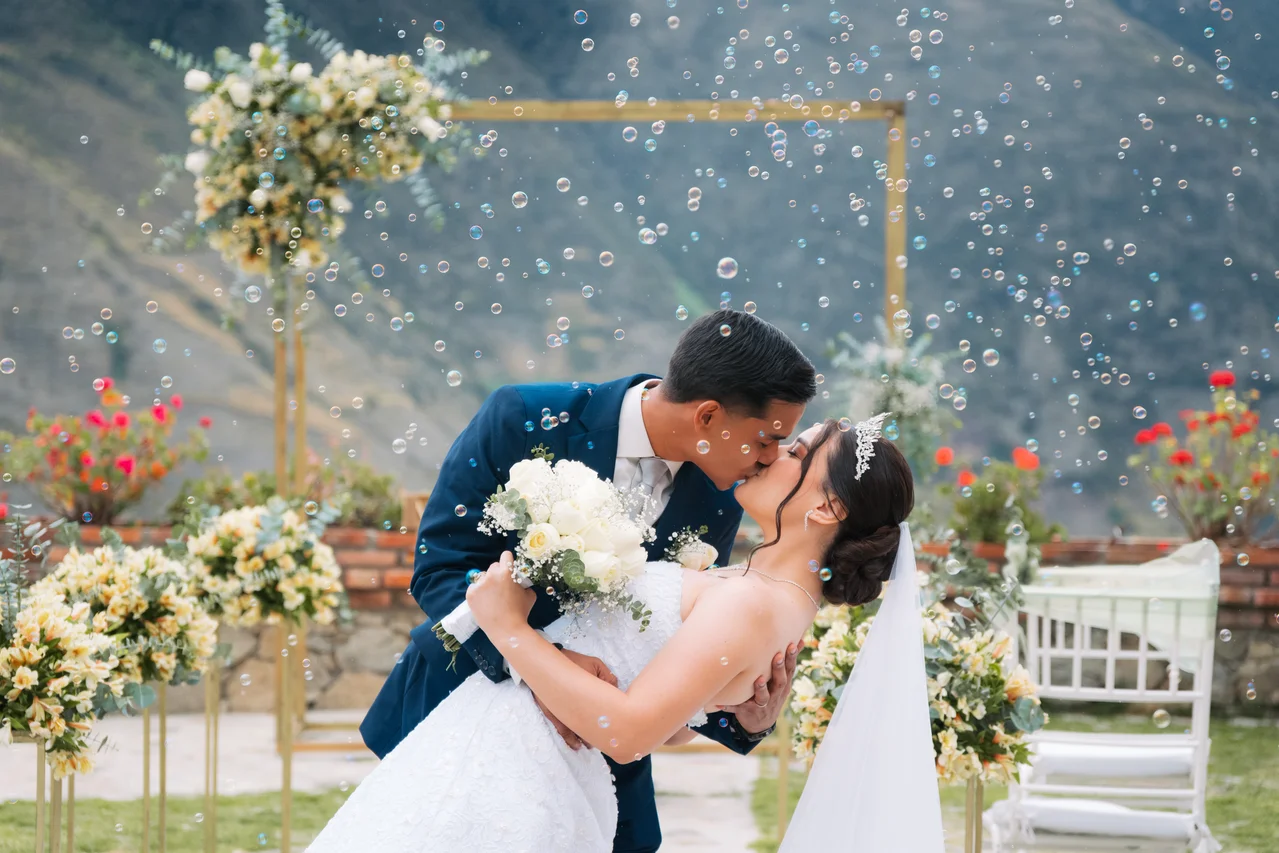 Beso de novios con inclinación y burbujas bajo arco floral, montaña al fondo