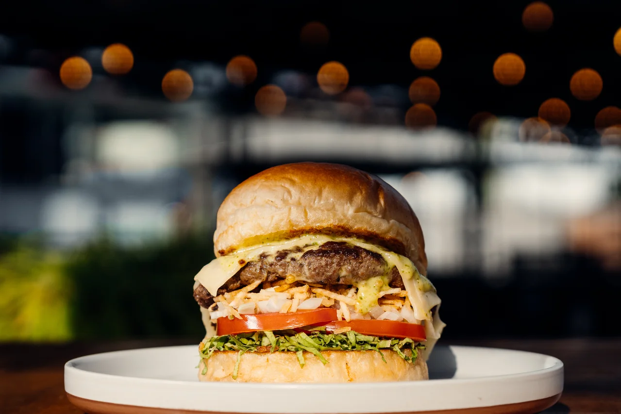 Hamburguesa centrada con sus capas nítidas, plato blanco y luces cálidas desenfocadas