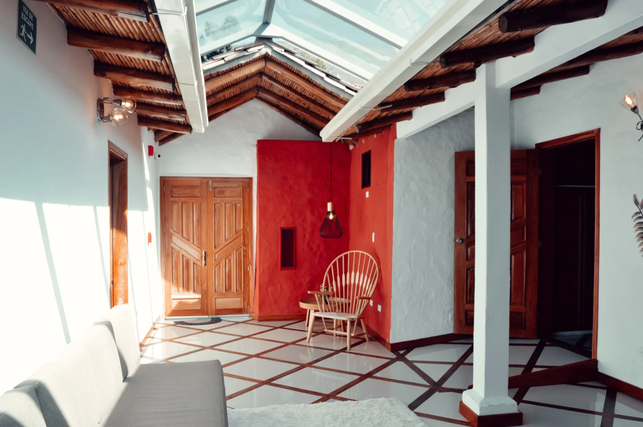 Atrio hotelero con techo de madera y vidrio, muro rojo central y columnas blancas