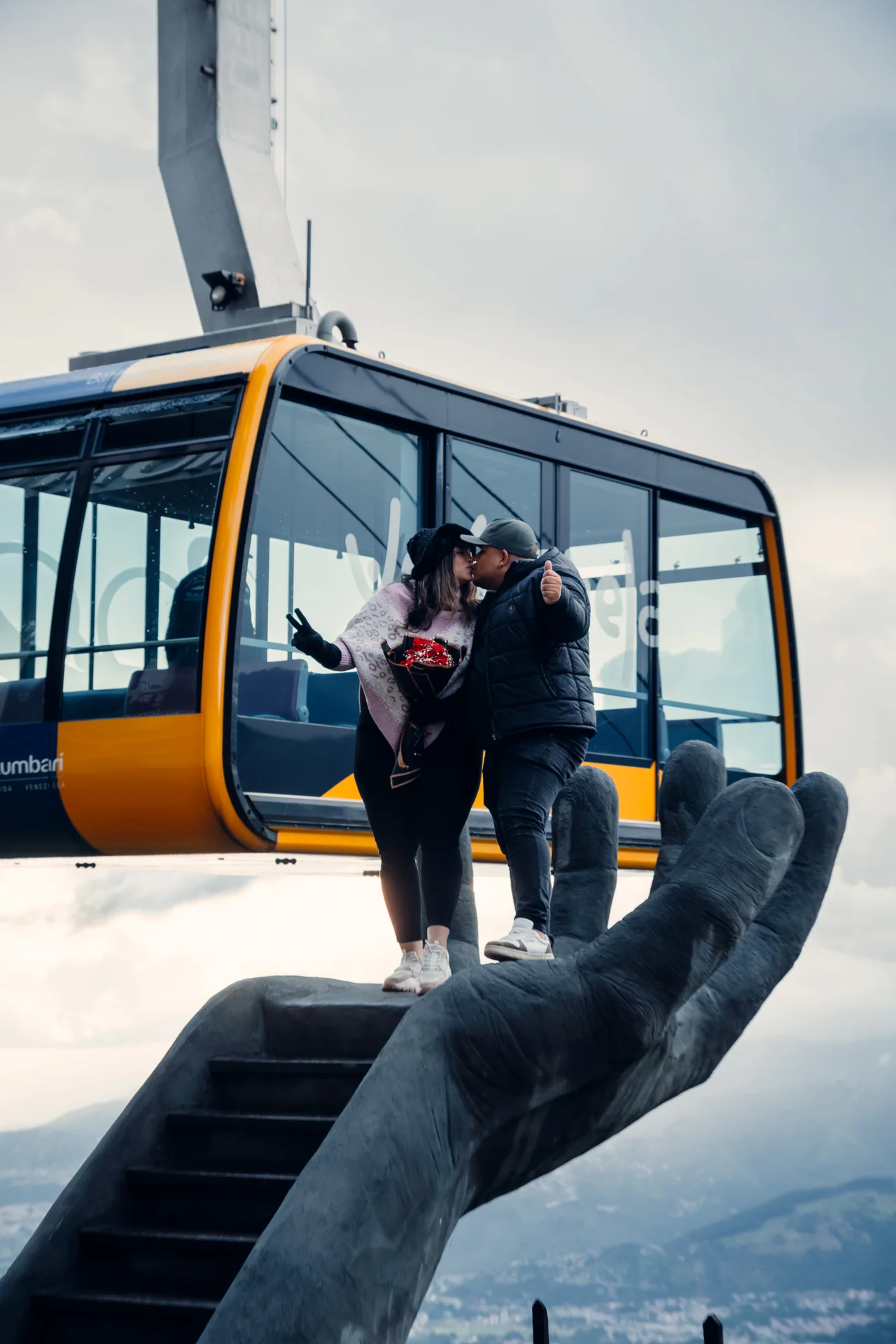 Pareja besándose sobre escultura de mano ante una cabina del teleférico