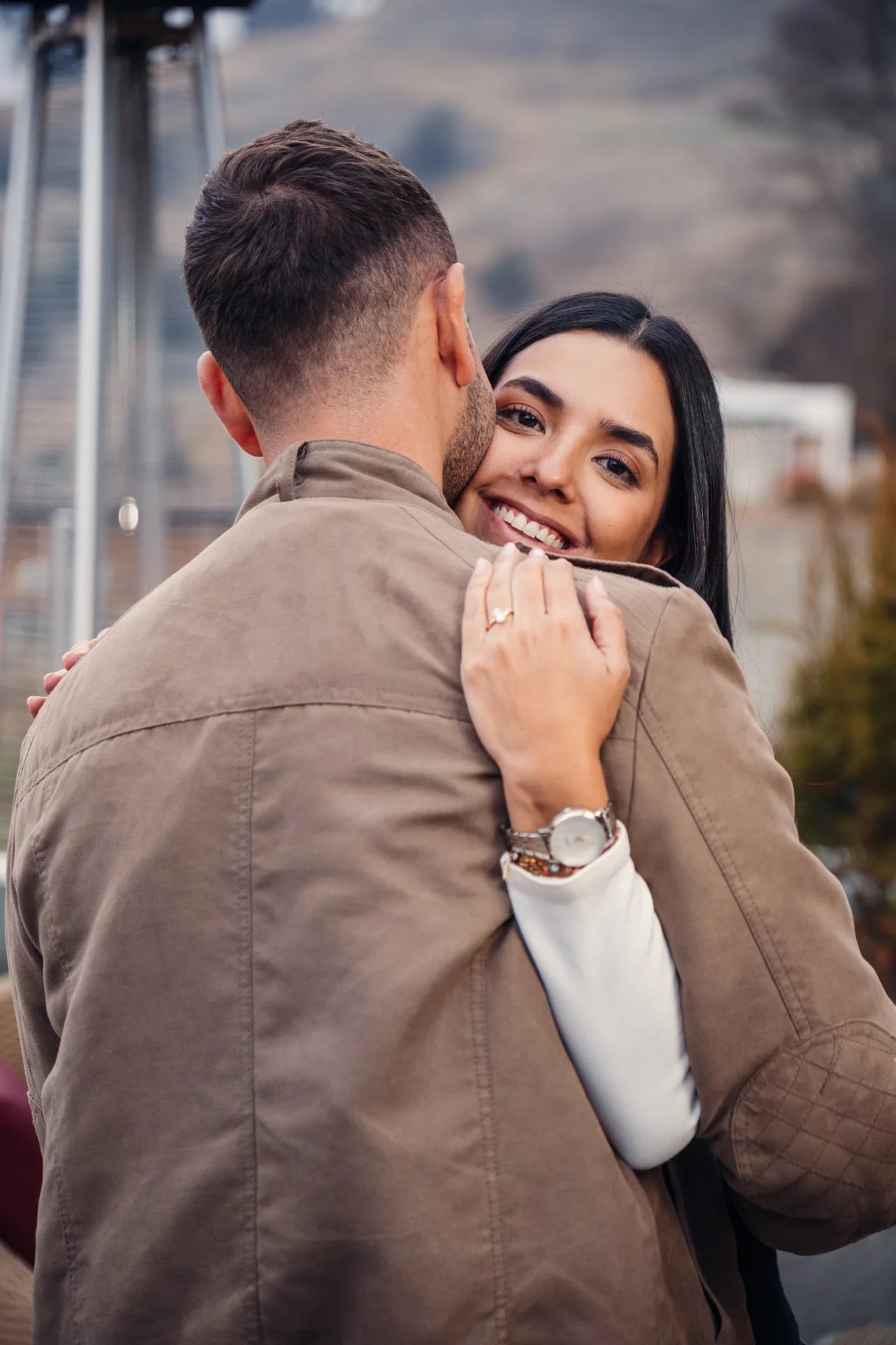 Retrato de pareja en montaña: él besa su mejilla y ella sonríe a cámara con el anillo visible